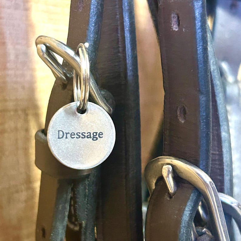 以下が含まれることがあります： Dressageという文字が刻印された円形の銀色の金属タグ。銀色の金属製バックルが付いたダークブラウンのレザーストラップに、二重リングで取り付けられています。タグは滑らかで平らで、中央に鮮明な黒いフォントで文字が記されています。