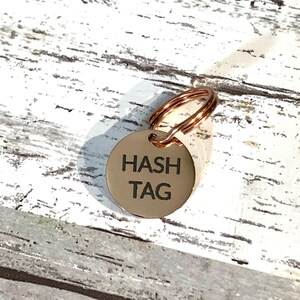以下が含まれることがあります： 太字の黒いサンセリフ体で「HASH TAG」と刻印された、円形のローズゴールドカラーの金属製ペット用タグ。タグの上部には小さな穴があり、同色のローズゴールドの二重リングが取り付けられています。