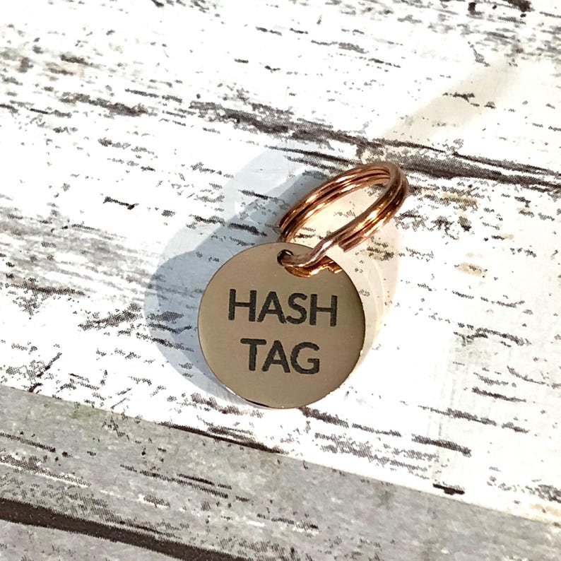 以下が含まれることがあります： 太字の黒いサンセリフ体で「HASH TAG」と刻印された、円形のローズゴールドカラーの金属製ペット用タグ。タグの上部には小さな穴があり、同色のローズゴールドの二重リングが取り付けられています。