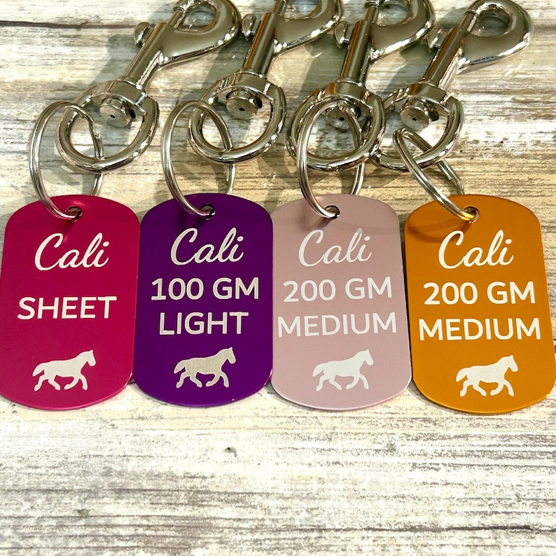 May include: Four metal horse tags with silver key rings. The tags are in different colors: pink, purple, light pink, and orange. The tags are engraved with the name "Cali" and different weight options: "Sheet", "100 GM Light", "200 GM Medium", and "200 GM Medium". Each tag has a small horse silhouette.