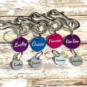 以下が含まれることがあります： Lucky、Grace、Princess、Roo Rooと刻印された紫、青、ピンク、濃い紫の円形タグが付いた4つの銀色の金属製回転式ロブスタークラスプ。その下には、同じ名前が刻印された4つの小さな銀色の円形タグが並んでいます。