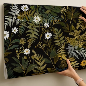 May include: A roll of wallpaper featuring a botanical design with white daisies, ferns, and other foliage in shades of green, gold, and gray against a black background. The wallpaper is being held up against a neutral wall.