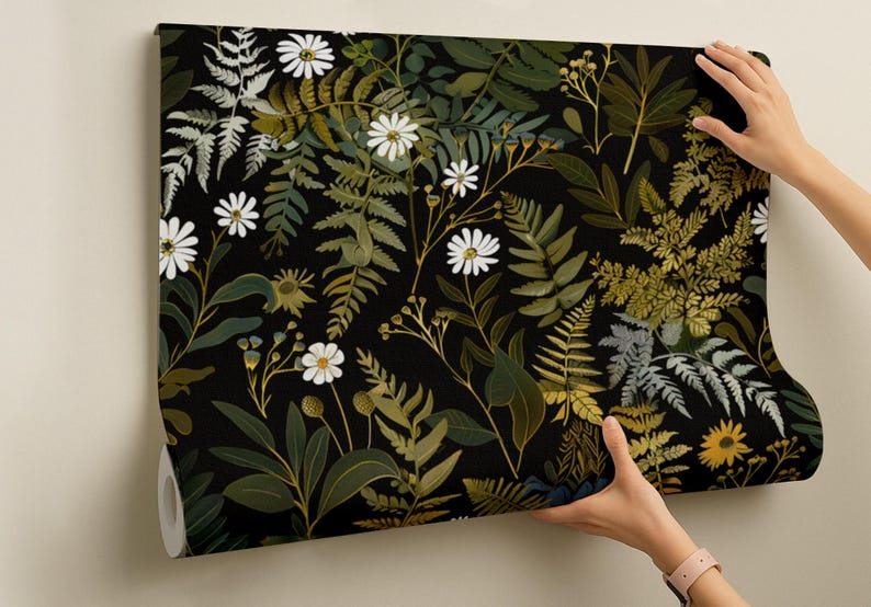 May include: A roll of wallpaper featuring a botanical design with white daisies, ferns, and other foliage in shades of green, gold, and gray against a black background. The wallpaper is being held up against a neutral wall.