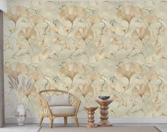 Op de afbeelding: Behang in beige met een herhaald patroon van gestileerde ginkgo bladeren en wijnstokken. De bladeren zijn in beige en bruine tinten, met subtiele schaduwen. Een rotan stoel, bijzettafels en een vaas met gedroogde planten staan voor het behang.