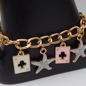 Pulsera única con dijes de estrella de la suerte con estrella gris con bordes dorados y dijes de jugador de cartas As de tréboles en rosa y blanco en una cadena de eslabones grandes de oro.