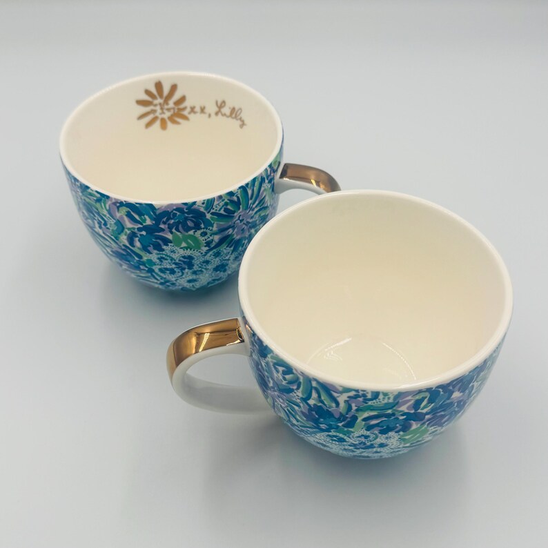 May include: Two white ceramic teacups with gold handles. The cups have a blue, green, and purple floral pattern on the outside. One cup has a gold flower and the text "xoxo, Lilly" inside.