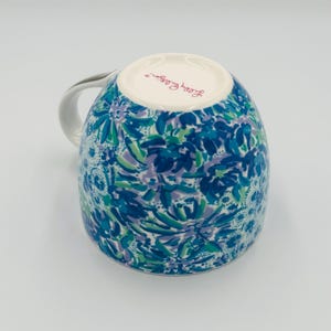 May include: A ceramic mug with a white handle and a floral pattern in shades of blue, green, and purple. The mug's interior is white, and the brand name "Lilly Pulitzer" is printed on the bottom.