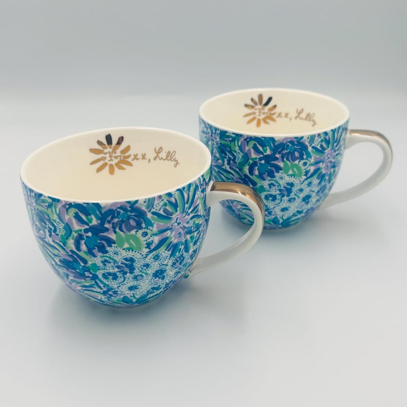 May include: Two ceramic teacups with a floral pattern in shades of blue, green, and purple. The cups have white interiors, gold-colored handles, and the text "xoxo, Lilly" inside. The cups are on a white surface.