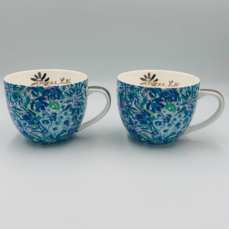 May include: Two ceramic mugs with a blue, green, and purple floral pattern. The mugs have white handles and a gold floral design with text inside. The mugs are on a white surface.