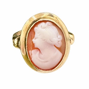 May include: An oval gold ring featuring a carved shell cameo. The cameo displays a light pink profile of a person against a darker peach-colored background, set within a polished gold frame with a smooth, rounded band on each side.
