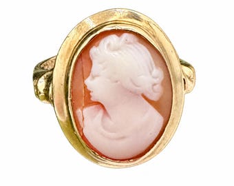 May include: An oval gold ring featuring a carved shell cameo. The cameo displays a light pink profile of a person against a darker peach-colored background, set within a polished gold frame with a smooth, rounded band on each side.