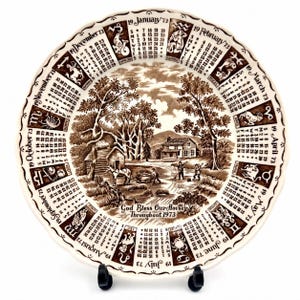 May include: A round ceramic decorative plate featuring a brown and white transferware print of a 1973 calendar. The border displays twelve months with zodiac symbols, while the center depicts a rural farm scene with the text God Bless Our House Throughout 1973.