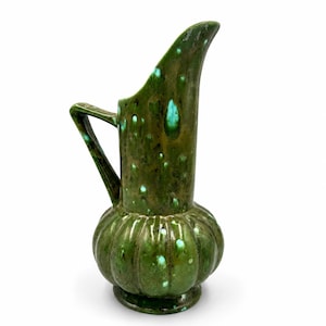May include: A ceramic pitcher featuring a deep olive green glaze with mottled turquoise and white speckles. The vessel has a bulbous, ribbed base, a tall cylindrical neck with a slanted rim, and a single angular handle attached to the side.