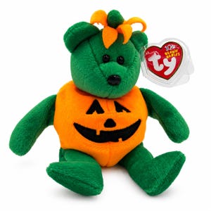 May include: A green plush bear wearing an orange pumpkin-shaped torso with a black stitched jack-o'-lantern face. The bear has orange fabric leaves on its head and a heart-shaped red Ty Beanie Babies tag encased in a clear plastic protector.