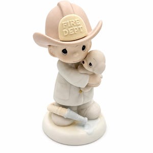 Peut inclure: Figurine en céramique d'un pompier tenant un petit animal. Le pompier porte un casque beige avec "FIRE DEPT" dessus. La figurine est principalement blanche et beige, avec un petit tuyau à ses pieds.
