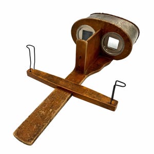 May include: Antique stereoscope viewer made of wood and metal. The viewer has two eye holes, a handle, and a crossbar for stability. The wood has a warm, aged tone, and the metal components are black.