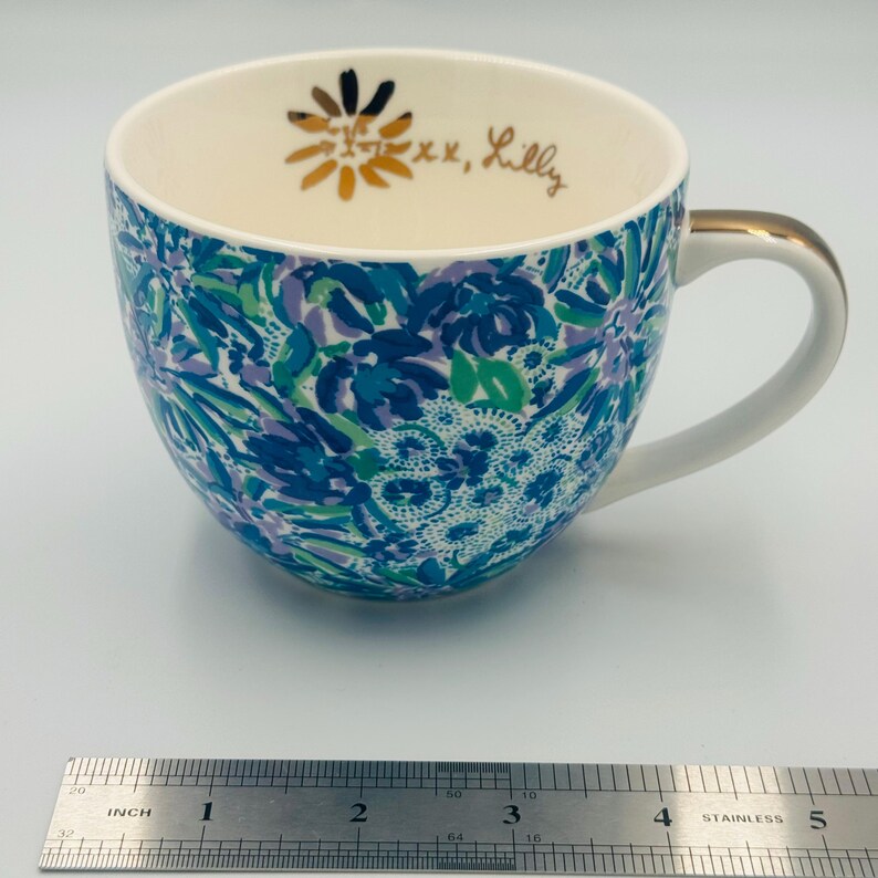 May include: A ceramic coffee mug with a white handle and gold trim. The mug features a blue, green, and purple floral pattern. The inside rim has gold text that reads "xx, Lilly" and a gold flower design. A ruler shows the mug is approximately 4 inches wide.