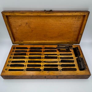 May include: An open wooden box containing a set of vintage letterpress printing type. The box is made of light brown wood and has a hinged lid. The type is arranged in rows and columns, with each letter in its own compartment.