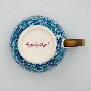 May include: A ceramic mug with a colorful floral pattern in shades of blue, green, and purple. The mug has a gold-colored handle and the words "Lilly Pulitzer" in red script on the bottom.