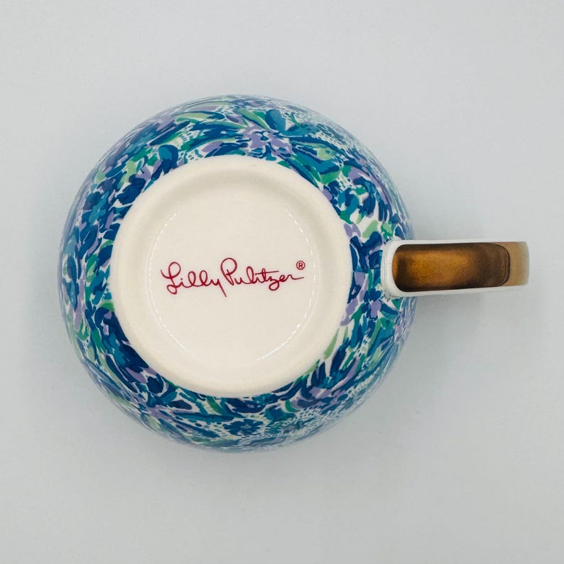 May include: A ceramic mug with a colorful floral pattern in shades of blue, green, and purple. The mug has a gold-colored handle and the words "Lilly Pulitzer" in red script on the bottom.