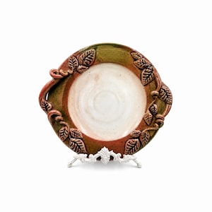 May include: A handcrafted ceramic plate with a white center, surrounded by a rim of green and reddish-brown hues. The plate is adorned with a leaf design and rests on a white decorative stand, showcasing a rustic aesthetic.