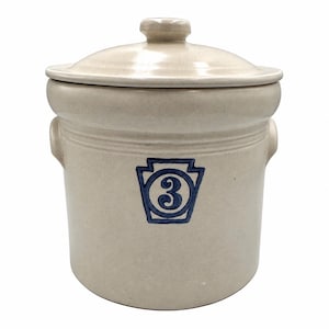 May include: A light beige stoneware crock with a matching lid and two side handles. The front features a blue stamped graphic of a keystone shape containing the number 3. The cylindrical container has a smooth finish and a wide rim.
