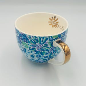 May include: A ceramic teacup with a floral pattern in shades of blue, green, and purple. The handle is gold-colored, and the interior is cream-colored. A gold floral design is on the inside rim.