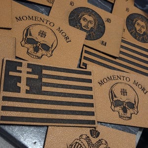 Può includere: Una collezione di toppe in pelle beige con vari disegni incisi in nero. Le toppe presentano immagini di un'aquila, un teschio con la scritta "Memento Mori", una bandiera e un ritratto di Gesù.