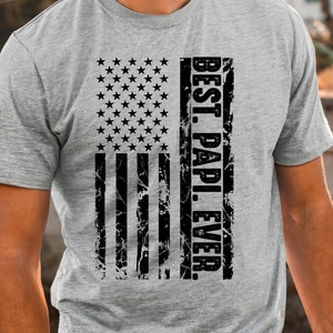 May include: A gray t-shirt with a distressed black and white American flag design. The text "BEST. PAPI. EVER." is printed vertically on the right side of the flag.