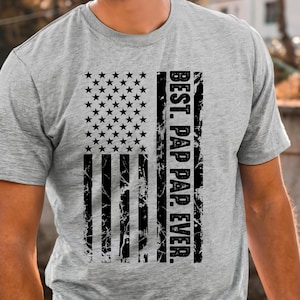 May include: A gray t-shirt with a distressed black and white American flag design. The text "BEST. PAP. PAP. EVER." is printed vertically on the right side of the flag.