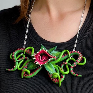 Könnte beinhalten: Eine grüne und rote Halskette mit einer großen, offenmündigen fleischfressenden Pflanze mit Tentakeln. Die Pflanze hat eine rosa Zunge und weiße Zähne.