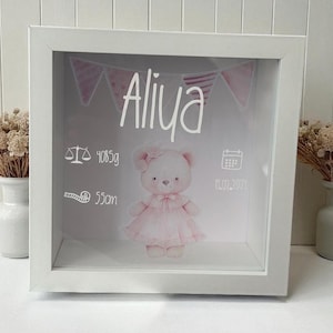 Könnte beinhalten: Ein weißer Schattenrahmen mit Glasfront. Im Inneren ist eine rosa Teddybär-Illustration unter dem Namen "Aliya" zentriert. Die Box enthält Details: Gewicht 4085g, Länge 55cm und Datum 14.02.2021.