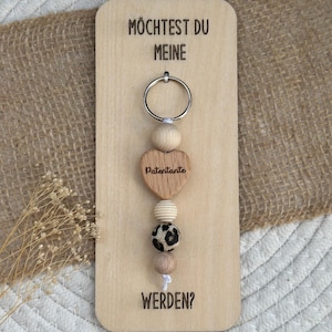 Könnte beinhalten: Ein Holzschlüsselanhänger mit einem herzförmigen Anhänger mit der Aufschrift "Patentante". Der Schlüsselanhänger hat eine Reihe von Holz- und gemusterten Perlen. Der Holzrücken trägt den Text "Möchtest du meine werden?"