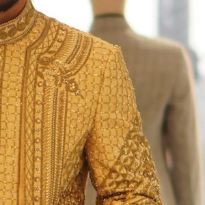 K&ouml;nnte beinhalten: Ein Mann tr&auml;gt eine goldene bestickte Jacke und eine passende Hose. Die Jacke hat einen Stehkragen und ein langes, flie&szlig;endes Design. Die Hose ist geschnitten und hat ein gerades Bein.