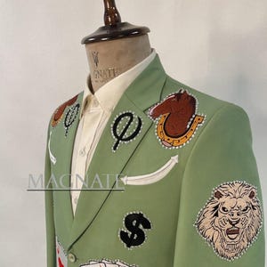 May include: Light green blazer with various embroidered patches, including playing cards, dice, a lion, and a dollar sign. The blazer has a white shirt underneath and the word "MAGNATE" is visible. The blazer is on a mannequin.