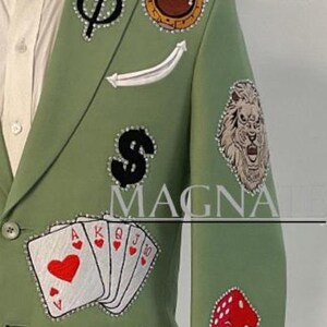 May include: Sage green blazer with various embroidered patches including playing cards, dice, a lion, and a chess set. The blazer has a white shirt underneath and is displayed on a mannequin.