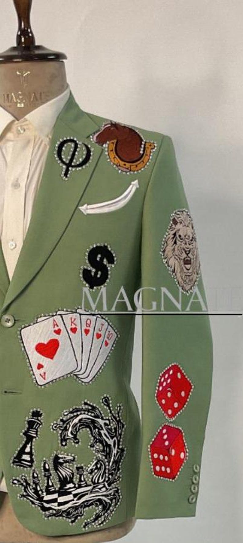 May include: Sage green blazer with various embroidered patches including playing cards, dice, a lion, and a chess set. The blazer has a white shirt underneath and is displayed on a mannequin.