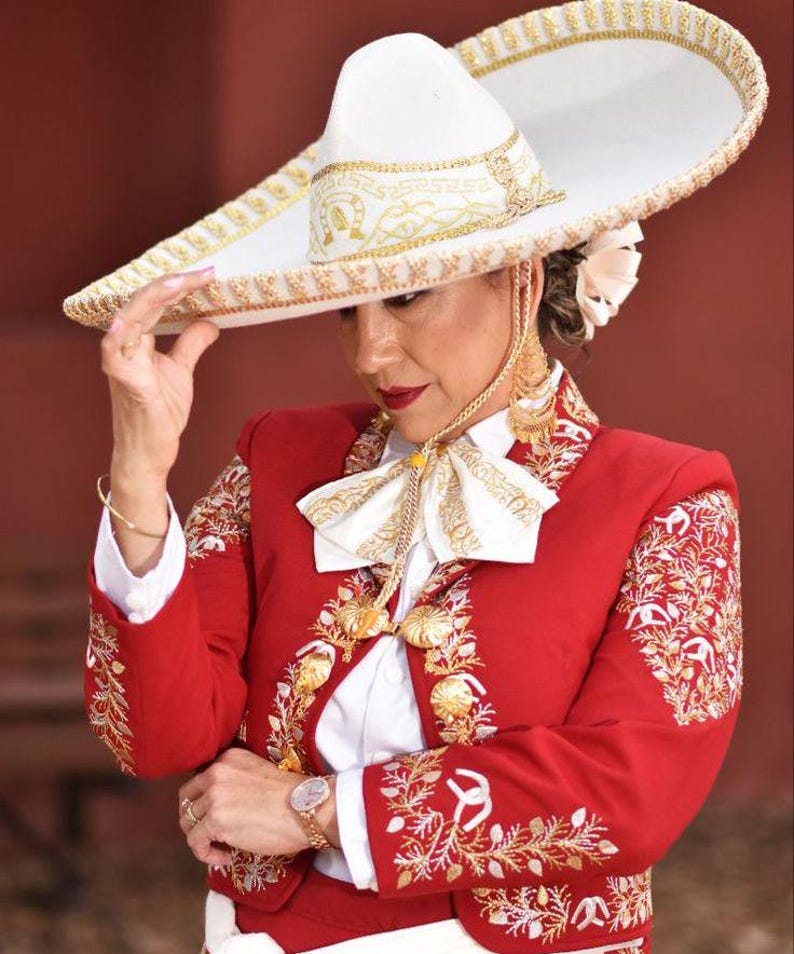May include: A woman wearing a red charro suit with gold embroidery and a white shirt with a bow tie. She is wearing a large white sombrero with gold trim and holding the brim of the hat with her left hand.