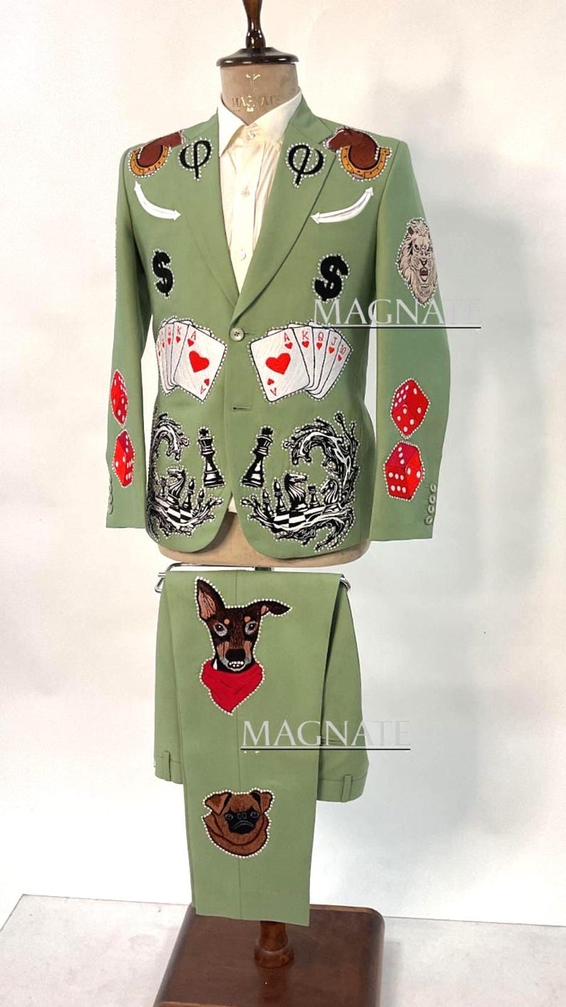May include: A sage green suit with a white dress shirt. The blazer features playing card suits, dice, and dog patches. The pants have dog patches. The suit is displayed on a mannequin.