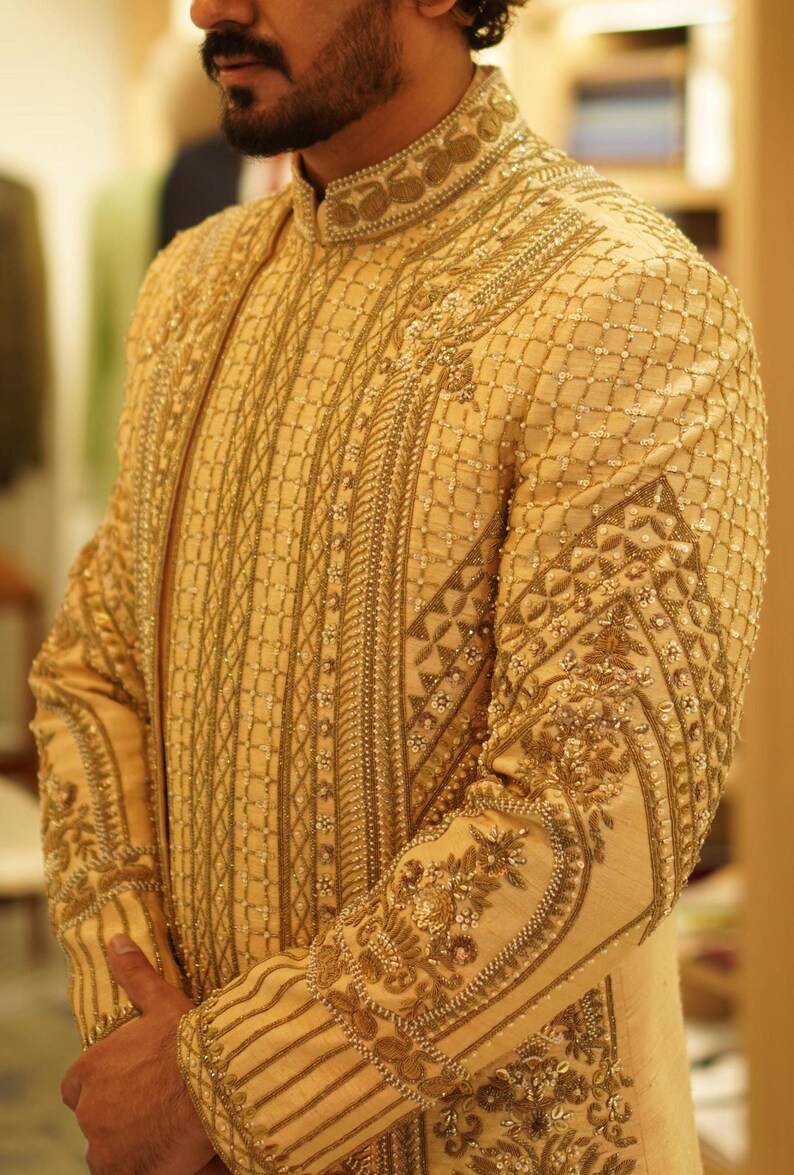 K&ouml;nnte beinhalten: Eine beige bestickte Jacke mit goldenen F&auml;den und Verzierungen. Die Jacke hat einen Stehkragen und lange &Auml;rmel.