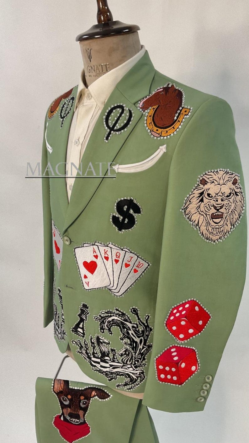 May include: Light green blazer with various embroidered patches, including playing cards, dice, a lion, and a dollar sign. The blazer has a white shirt underneath and the word "MAGNATE" is visible. The blazer is on a mannequin.