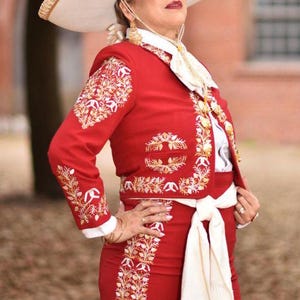May include: A red charro suit with gold embroidery and a white sash. The suit includes a jacket with long sleeves and a skirt. The person is wearing a white shirt with a lace collar and a white hat.