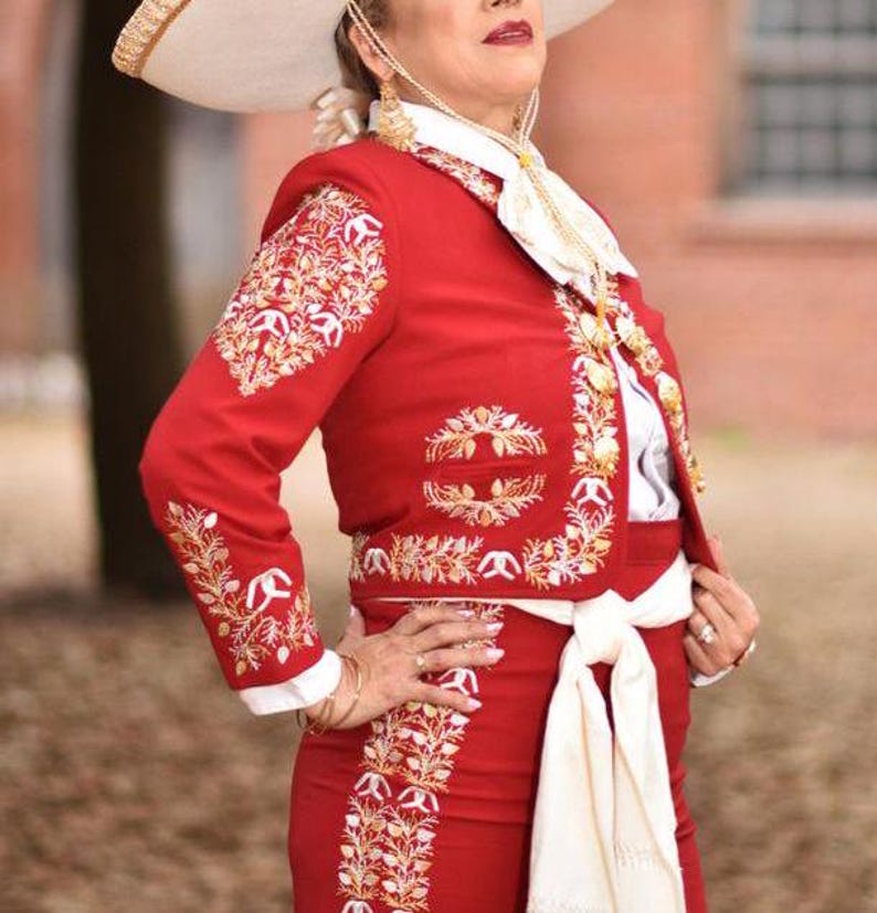 May include: A red charro suit with gold embroidery and a white sash. The suit includes a jacket with long sleeves and a skirt. The person is wearing a white shirt with a lace collar and a white hat.