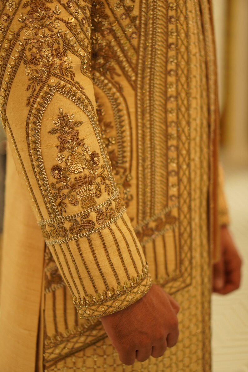 K&ouml;nnte beinhalten: Ein golden besticktes Kleidungsst&uuml;ck mit komplizierten floralen und geometrischen Mustern. Der Stoff hat einen dezenten Glanz und ist mit zarten Perlen verziert.
