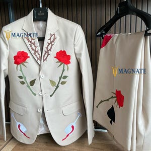 Könnte beinhalten: Ein beiger Anzugsakko und eine passende Hose mit gestickten Rosen und Weingläsern. Das Sakko hat ein braunes Dornenmuster am Revers. Die Hose hat ein schwarzes Pik-Muster. Der Anzug ist mit "MAGNATE" gekennzeichnet.