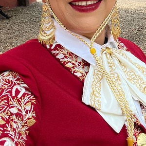 May include: A red embroidered jacket with a white embroidered collar and a gold chain with a yellow bead. The jacket has a floral pattern and is worn by a person with a gold earring.