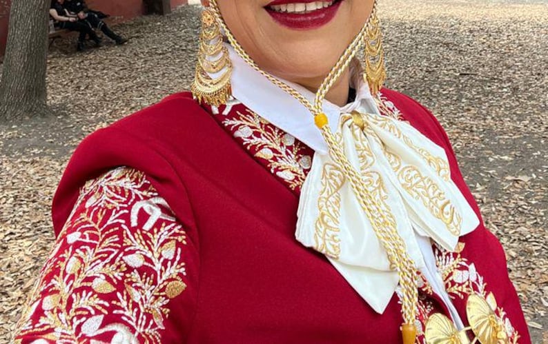 May include: A red embroidered jacket with a white embroidered collar and a gold chain with a yellow bead. The jacket has a floral pattern and is worn by a person with a gold earring.