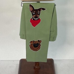 May include: Light green pants with embroidered dog portraits. One dog is a brown and black breed wearing a red bandana, and the other is a brown pug. The pants are displayed on a wooden hanger.