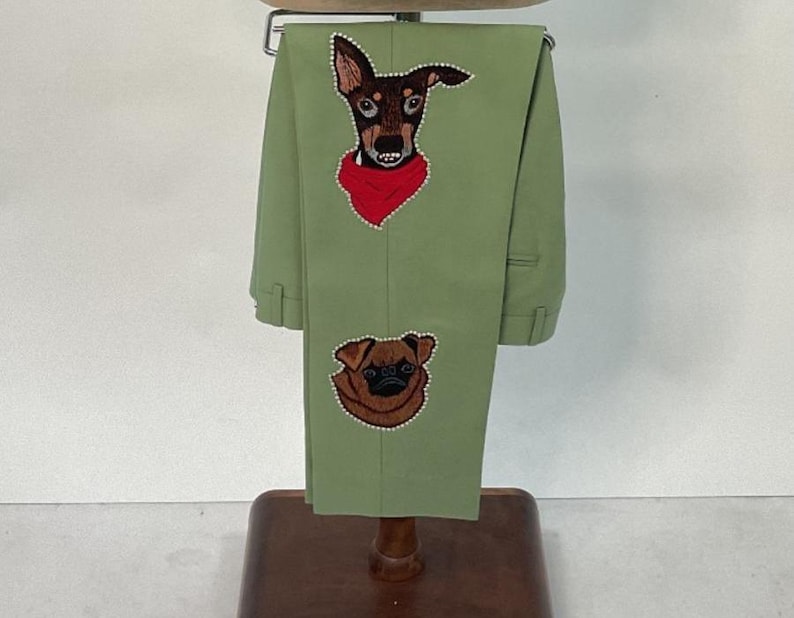 May include: Light green pants with embroidered dog portraits. One dog is a brown and black breed wearing a red bandana, and the other is a brown pug. The pants are displayed on a wooden hanger.