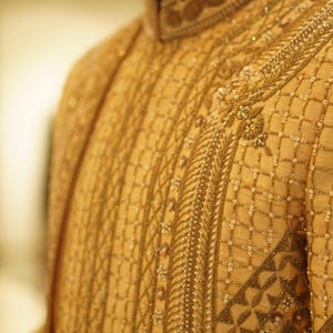 K&ouml;nnte beinhalten: Nahaufnahme eines goldenen bestickten Kleidungsst&uuml;cks mit komplizierten Mustern und Perlen. Der Stoff ist hellbeige und die Stickerei ist mit einem dunkleren goldenen Faden ausgef&uuml;hrt.